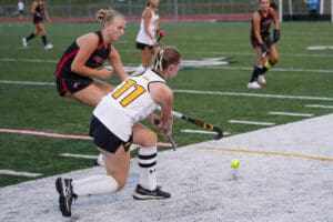 Field hockey players in action, player in white and black uniform about to hit the ball on a green turf field.
