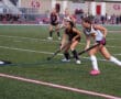 Two field hockey players chase the ball during a competitive match on a grassy field.