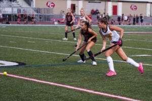 Two field hockey players chase the ball during a competitive match on a grassy field.