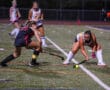 Girls field hockey match at night, player in black defends against player in white and yellow near goal.