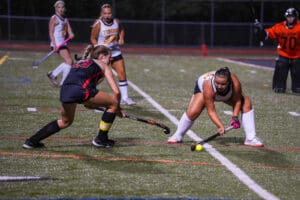 Girls field hockey match at night, player in black defends against player in white and yellow near goal.