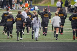 Field hockey team, wearing North Allegheny jackets, running on a sports field, sticks in hand.
