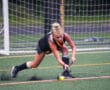 Field hockey player focused, striking a ball on artificial turf near the goal post.