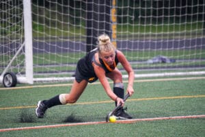 Field hockey player focused, striking a ball on artificial turf near the goal post.