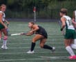 Field hockey player in black uniform hits the ball, facing opponents in green on a turf field.