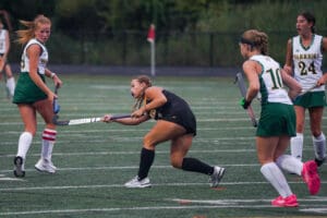 Field hockey player in black uniform hits the ball, facing opponents in green on a turf field.