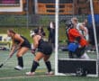 Field hockey players in black uniforms near the goal, goalie in orange, during an intense match in the rain.