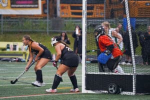 Field hockey players in black uniforms near the goal, goalie in orange, during an intense match in the rain.
