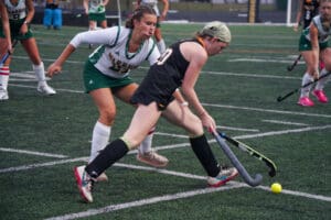 Two female field hockey players competing for the ball during a match on a green turf field.