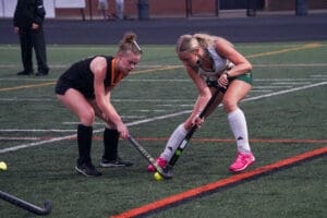 Two female field hockey players competing for the ball on a turf field during a match.