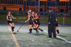 Field hockey team celebrating a victory on the field with coach nearby, school buses in the background.
