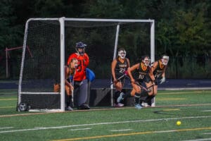 Field hockey players in action, defending a goal during an intense match on a green turf field.