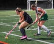 Two female field hockey players competing on a turf field at night.