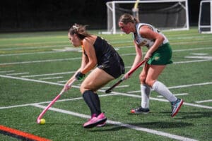 Two female field hockey players competing on a turf field at night.