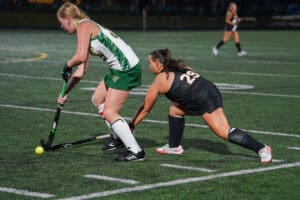 Two female field hockey players competing for the ball on a green turf field.