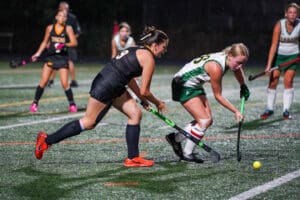Field hockey players competing on a turf field at night, focusing on the action around the ball.