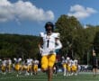 Football player in yellow and white uniform runs on field with team warming up under a blue sky.