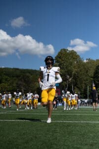 Football player in yellow and white uniform runs on field with team warming up under a blue sky.