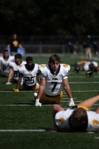 Football players stretch on a field in team practice, wearing yellow and white uniforms.