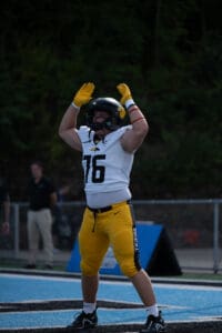 Football player in yellow pants and white jersey raises arms on field.
