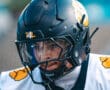 Football player in black helmet and white jersey with yellow accents, focused before the game.