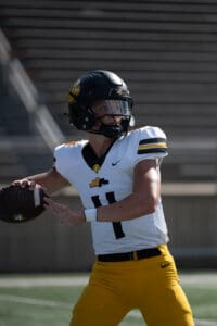 Football player wearing a helmet prepares to throw, dressed in a white jersey and yellow pants on a field.