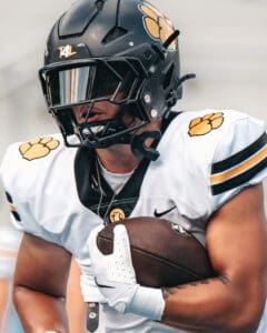 Football player in black helmet and white uniform clutching a football during a game, showcasing team spirit and focus.