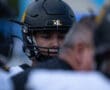 A football player in a black helmet focused before a game, surrounded by teammates in blurred foreground.
