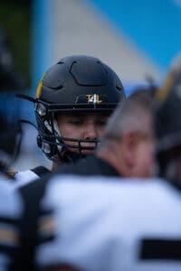 A football player in a black helmet focused before a game, surrounded by teammates in blurred foreground.