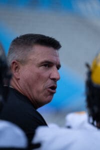 Man speaking passionately in a sports setting, wearing a black shirt, with blue background.