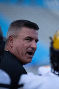 Man in a sports setting, speaking passionately, wearing a black shirt with blurred bleachers in the background.