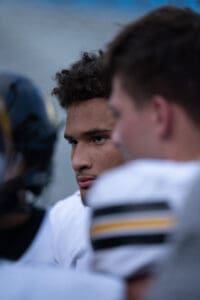 Football player focused on the game, wearing a white jersey with black and yellow accents, during a huddle.