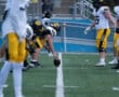 Football players prepare for snap during game on field, wearing black and yellow uniforms.