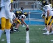 Football team lines up for a play on the field, players in black helmets and yellow pants focus on the upcoming snap.