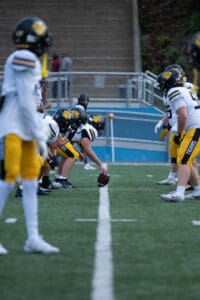 Football team lines up for a play on the field, players in black helmets and yellow pants focus on the upcoming snap.