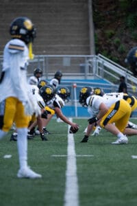 Football teams in yellow and white uniforms face off on the field, ready to start a play.