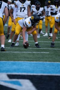 Football team preparing for snap on the field, players in yellow and white uniforms, focus on center holding ball.