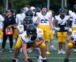 Football players in formation on the field during practice. Players wear white jerseys and yellow pants.