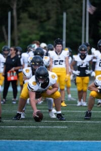 Football players in formation on the field during practice. Players wear white jerseys and yellow pants.