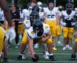 Football players practice snap drills in yellow and white uniforms on a field with trees in the background.