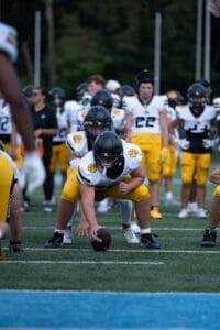 Football players practice snap drills in yellow and white uniforms on a field with trees in the background.