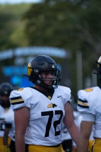 Football player in white and yellow uniform, number 72, wearing a black helmet, standing on the field.