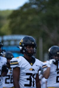 Football player in black helmet and white uniform, number 31, on the field with teammates in background.