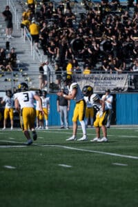 High school football game, players in yellow preparing for a snap, crowd and band in the stadium background.