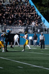 High school football game action with marching band in the stands, featuring a quarterback passing the ball.