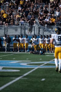 Football player running as opposing team watches from bench, spectators in stands, sunny day, competitive match atmosphere.