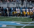 High school football game with players in action and spectators in the stands. Players in yellow pants lined up.