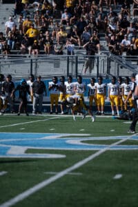 High school football game with players in action and spectators in the stands. Players in yellow pants lined up.
