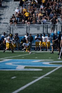 Football game action with players in yellow and blue uniforms, spectators in stands, daylight match.