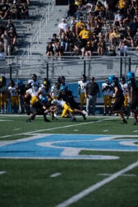 Football game action with players tackling on field, spectators in stands, vibrant team uniforms, sunny day.